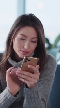 Sosyal medya uygulaması için cep telefonu kullanan ve oturma odasında kahve içen oldukça beyaz, samimi, kazaklı bir bayan..