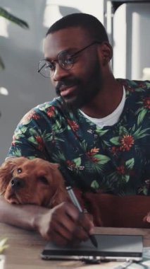 Çiçekli tişörtlü bir adam, yanında Golden Retriever 'la tablete resim çiziyor. İş hayatı dengesini vurguluyor..