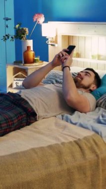Bir adam rahat bir şekilde yatağında uzanıyor, akıllı telefonuyla rahat ve modern bir yatak odası boşluğunda.