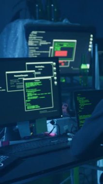 Loş bir odada, bir kapüşonlu bilgisayar başında çalışıyor. Monitörler, üst üste gelen yeşil metni dikkatle yazarken gösteriyor.