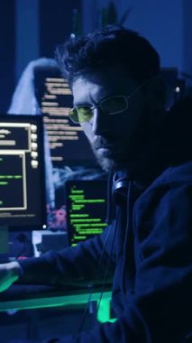 Loş bir odada, insanlar perdeleri analiz ederken kapüşonlu bir adam iki monitörü incelerken, güneş gözlüklü bir diğeri de gözetlemeye odaklanıyor.