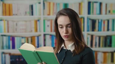 Kendini işine adamış ve odaklanmış genç bir kadın samimi bir kütüphane atmosferinde kitap okumaya dalmış durumda.