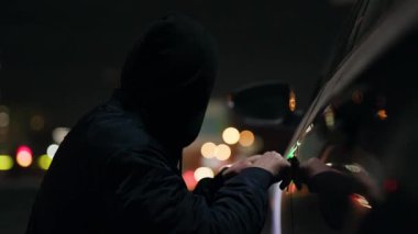 Gizemli bir kukuletalı şahıs karanlık gece gökyüzünün örtüsü altında park halindeki bir arabaya girmeye çalışıyor.