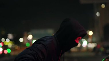 Gizlenmiş bir figür, loş bir kentsel ortamda gölgelerle örtülü duruyor ve derin bir entrika hissi uyandırıyor.