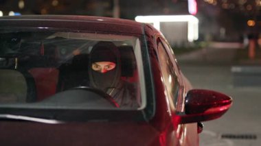 Gizemli, maskeli bir şahıs, park halindeki bir araçta sessizce oturuyor ve gözle görülür bir entrika ve gerilim hissi yaratıyor.