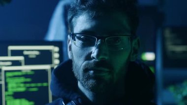 Gözlüklü odaklanmış bir kişi, kod ve programlama gösteren dijital ekranlarla çevrili karanlık bir alandadır.