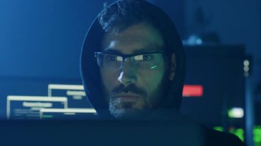 Kapüşonlu bir giysi giyen genç bir programcı, loş bir ortamda bir bilgisayarın üzerinde çalışıyor.
