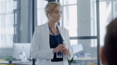 Beyaz ceketli profesyonel bir kadın ofiste güven içinde duruyor, iş arkadaşlarıyla yakınlaşıyor ve dinamik, aydınlık bir ortamda işbirliğini teşvik ediyor.