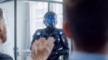 Modern bir ofiste, bir adam bir robotla konuşur, işbirliği ve dostluk göstererek coşkuyla el çırpar, ortaklığı sembolize eder.