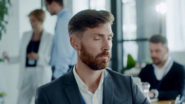 Sakallı bir erkek sunucu modern bir ofiste seyirciyle iletişim ve liderlik becerileri sergileyerek takım çalışmasını ön plana çıkarıyor.