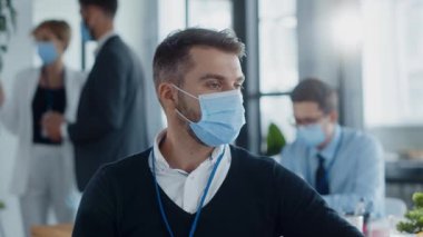 Koruyucu yüz maskesi takan bir adam ofis ortamında bir masada oturuyor, muhtemelen işle meşgul ya da önemli görevleri çalışıyor.