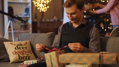 Siyah yelekli ve kareli gömlekli genç yakışıklı adam kanepeye oturmuş ve birbirine aşık mutlu bir çiftin arka planında Noel hediyesini gözden geçiriyor. Gülümsüyor. Kapalı.