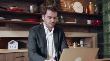 Resmi iş kıyafeti giymiş bir adam şık bir mutfak ortamında dizüstü bilgisayarına konsantre oluyor.