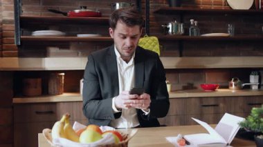 Takım elbiseli bir adam şık bir mutfak masasında akıllı telefonuyla modern yaşam tarzını yansıtıyor..