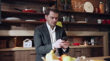 Profesyonel bir adam şık ve modern bir mutfak ortamında otururken akıllı telefonuyla aktif olarak meşgul oluyor.