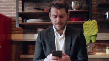 Akıllı takım elbiseli odaklanmış bir işadamı şık bir mutfak ortamında akıllı telefonuyla mesajlaşıyor.