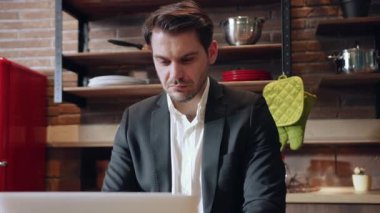 Özel dikim takım elbise giymiş kendini işine adamış bir adam şık bir mutfakta şık bir dizüstü bilgisayar üzerinde çalışıyor.