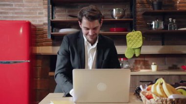 Şık bir ceket giyen odaklanmış bir adam modern bir mutfak düzeninde otururken özenle bir dizüstü bilgisayar üzerinde çalışıyor.