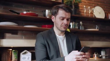 Samimi ve şık bir mutfakta, genç bir adam telefonunu kontrol ederken endişeli ya da üzgün görünüyor.
