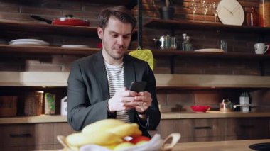 Modern bir mutfakta taze meyve dolu dururken şık bir adam akıllı telefonuyla meşgul olur.