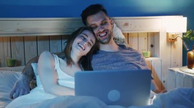 Neşeli bir çift yatağında rahat bir şekilde kıvrılıp dizüstü bilgisayarlarını kullanırken rahat bir film gecesinin tadını çıkarıyor.