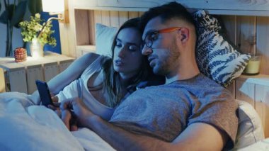 Sevgi dolu bir çift, gece boyunca mutlu mesut akıllı telefon kullanarak rahat yataklarında vakit geçirmenin keyfini çıkarıyor.