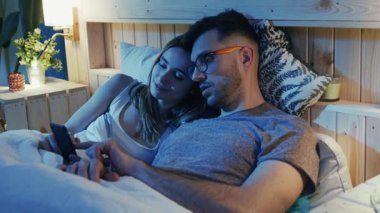 Samimi bir çift yatakta birbirine sokulmuş, telefon kullanırken sakin ve sessiz bir anın tadını çıkarıyorlar.