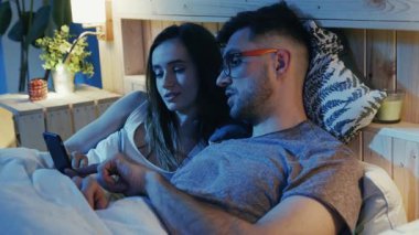 Sıcak ve rahat sahnelerde bir çift, akıllı telefon kullanırken yatakta keyifle vakit geçiriyorlar.