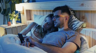 Bir çift yatakta rahat bir şekilde dinleniyor, akıllı telefona bakarken sıcak ve samimi bir anı paylaşıyor.