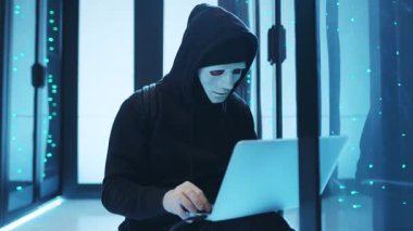 Maskeli bir hacker, ileri teknoloji ürünü bir sunucu odasında yüksek teknolojili bir dizüstü bilgisayarın başında çok sıkı çalışıyor.