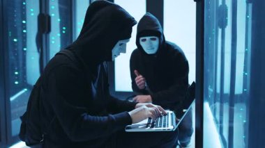 İki maskeli şahıs, gelişmiş bir sunucu içindeki yüksek teknolojili bir dizüstü bilgisayarın üzerinde çalışıyor. Siber entrikalar yayıyorlar.