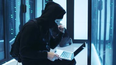 Kapüşonlu bir figür, loş bir veri merkezindeki sistemleri hackliyor ve görmezden gelemeyeceğimiz ciddi siber güvenlik risklerini vurguluyor.