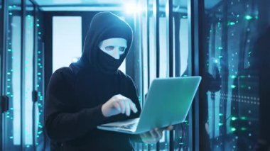 Gizemli maskeli bir şahıs yüksek teknolojili bir veri merkezinde siber aktivite gösteren parlak bir dizüstü bilgisayar kullanıyor.