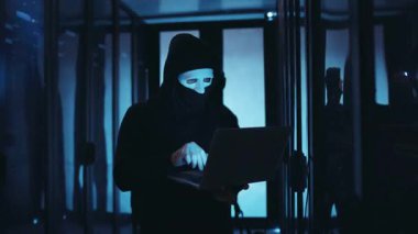 Maskeli bir başlıklı şahıs, hassas bir sunucu odasını bilgisayarla hackliyor ve günümüzde siber tehditlerin altını çiziyor.