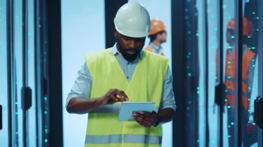 Çağdaş bir veri merkezinde çelik yelek ve kask giyen bir inşaat işçisi tablet kullanıyor.