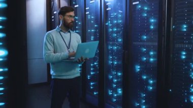 Bir sunucu odasında, bir adam dizüstü bilgisayarda çalışır, önemli görevlerle meşgul olur. Sahne modern teknolojiyi ve veri işlemenin önemini vurguluyor
