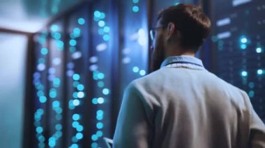 Bir sunucu odasında, bir adam dijital işlemleri yönetir, sunucuları yönetir ve telefonunu kullanır, tabletini kontrol eder ve performansını gözlemler.