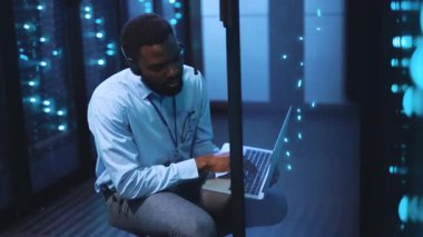 Odaklanmış bir adam sunucularla çevrili bir dizüstü bilgisayarla sunucu odasında çalışıyor. Verileri yönetirken kulaklıkları ayarlıyor, teknolojik yoğunluğu gösteriyor.