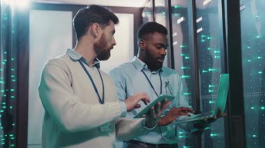 Farklı sakallı iki adam, teknoloji ve cihazlarla dolu bir sunucu odasındaki tablete dikkatle bakıyor.