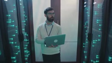 Sunucu odasında çalışan bir adam dizüstü bilgisayarındaki verileri sunucuların arasında yönlendiriyor, teknoloji ve uzmanlığı vurguluyor.