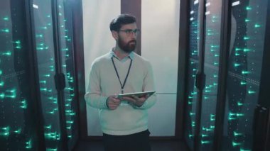 Bir adam şu anda sunucu odasında elinde tablet bir cihaz tutarken ve etrafındaki karmaşık teknolojiyi gözlemlerken...