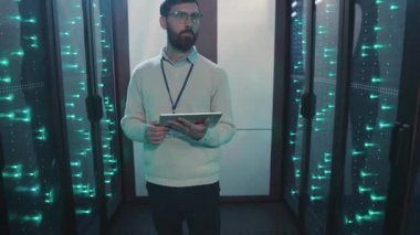 Bir IT uzmanı sunucularla dolu yüksek teknoloji sunucu odasındaki bir tabletin verilerini inceleyerek, modern teknoloji analizinde veri analizinin önemini vurguluyor.