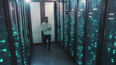 Yüksek teknolojili sunucu odasında, bir adam veri analiz etmek ve sistemleri izlemek için tablet kullanır. Kritik altyapı yönetiminde uzmanlığını gösterir.