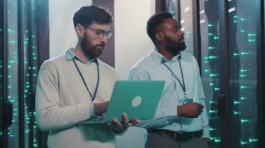 Yüksek teknolojili bir sunucu odasında, iki profesyonel bir dizüstü bilgisayardaki verileri analiz eder, sunucular ve yanıp sönen ışıklar arasında takım çalışması ve yenilikleri somutlaştırır.