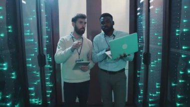 Bir veri merkezindeki iki adam yüksek teknoloji sunucu rafları etrafında işbirliği yapıyor, verileri analiz ederken uzmanlık gösteriyorlar, teknolojilerin rolünü vurguluyorlar.