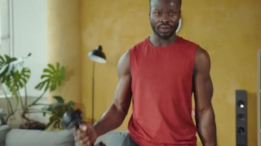 Bir fitness meraklısı gücünü parlak bir oturma odasında gösterir. Dambılları kaldırıyor, etkili ev egzersizleri tekniklerini gösteriyor.