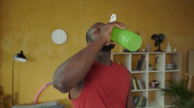 Konforlu bir oturma odasında yeşil bir şişeden su içen bir adam, canlı kırmızı bir atlet giymiş, neşeli bir şekilde hula çemberi tutuyor.