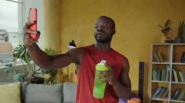 Bir adam rahat bir oturma odasında selfie çekiyor, bir elinde su şişesi, diğer elinde akıllı telefonu, sıradan bir titreşim uyandırıyor.