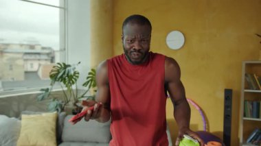 Rahat bir oturma odasında genç bir adam cep telefonunu kullanır, mesajları kontrol eder, suyla su içer ve kulaklıkla müzik dinler.