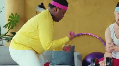 Arkadaşlar evde eğlenceli bir fitness seansının keyfini çıkarırken, diğerleri hula hoop kullanarak takım çalışmasını desteklerken, sarı gömlekli biri pembe halterler giyiyor.